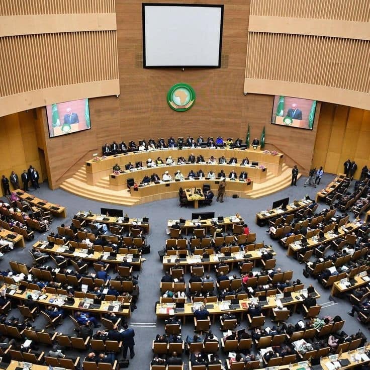 African Union Assembly in session in the plenary hall at AU headquarters in Addis Ababa, with AU flags beside the podium
